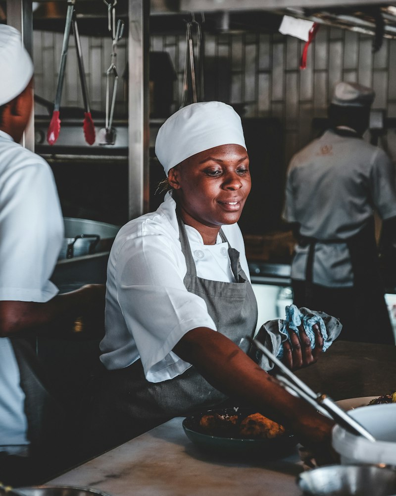 A cook in a white cap and apron plates food on a busy restaurant line
